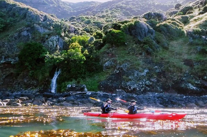 Two kayakers in a red kayak paddling below a small waterfall on a rocky, bush-covered shore